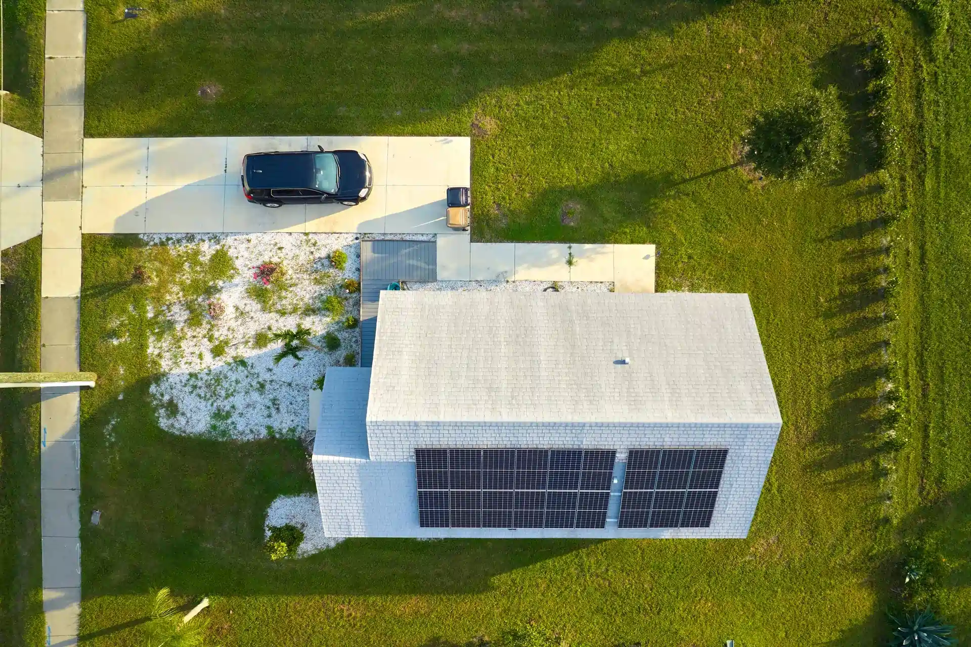 Letecký pohľad na rodinný dom so solárnymi panelmi na streche, s autom na príjazdovej ceste a zelenou záhradou okolo, znázorňujúci fotovoltiku pre domácnosti