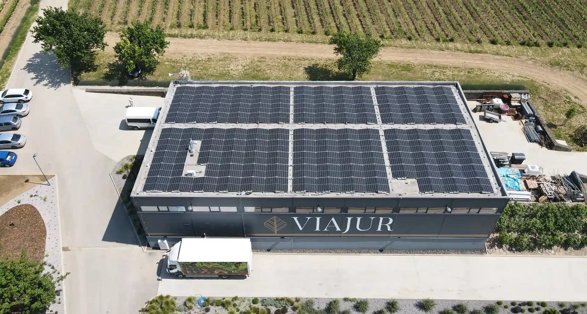 Letecký pohľad na priemyselnú budovu s rozsiahlou inštaláciou solárnych panelov na streche, obklopenú poľami a stromami, znázorňujúca fotovoltiku pre firmy