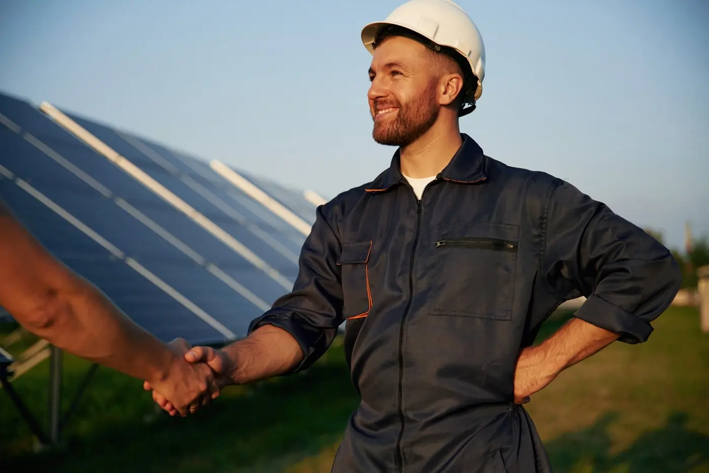 Usmievajúci sa technik v bielej ochrannej prilbe a tmavom pracovnom obleku stojaci pri solárnych paneloch, reprezentujúci inovácie a technologický pokrok v obnoviteľnej energii, Správa energetických zariadení od green energy