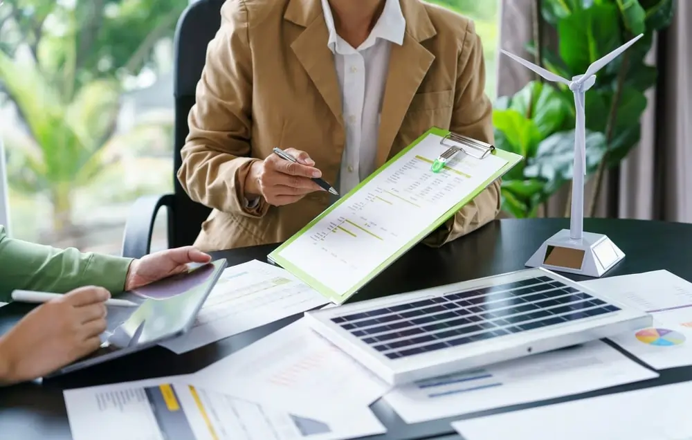 Žena v béžovom saku diskutuje nad projektovými dokumentmi a grafmi s laptopom na stole, reprezentujúca návrh technológie a profesionálne plánovanie