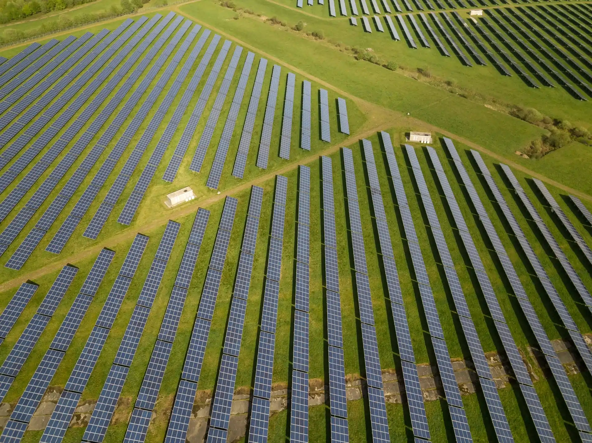Letecký pohľad na rozsiahlu solárnu elektráreň s radmi solárnych panelov usporiadanými na zelenej lúke, zobrazujúca veľkoplošnú inštaláciu obnoviteľnej energie